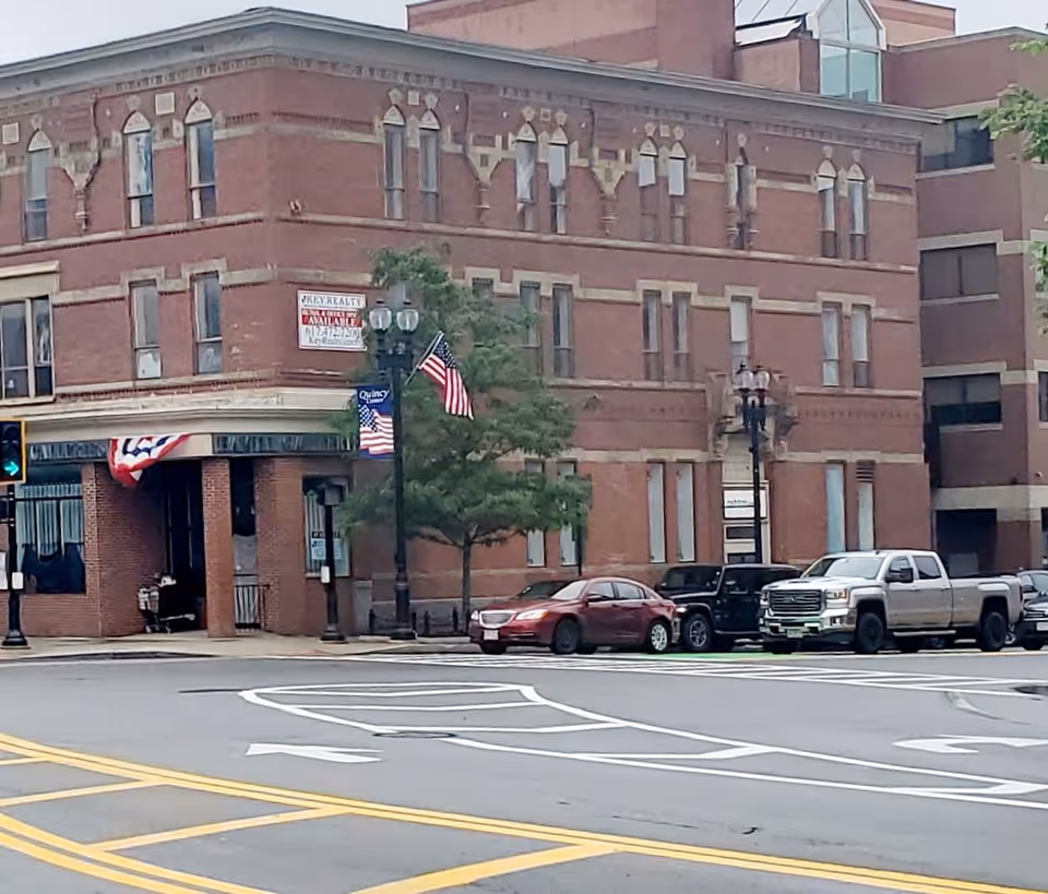 Brick building on a street corner with parked cars and American flags.