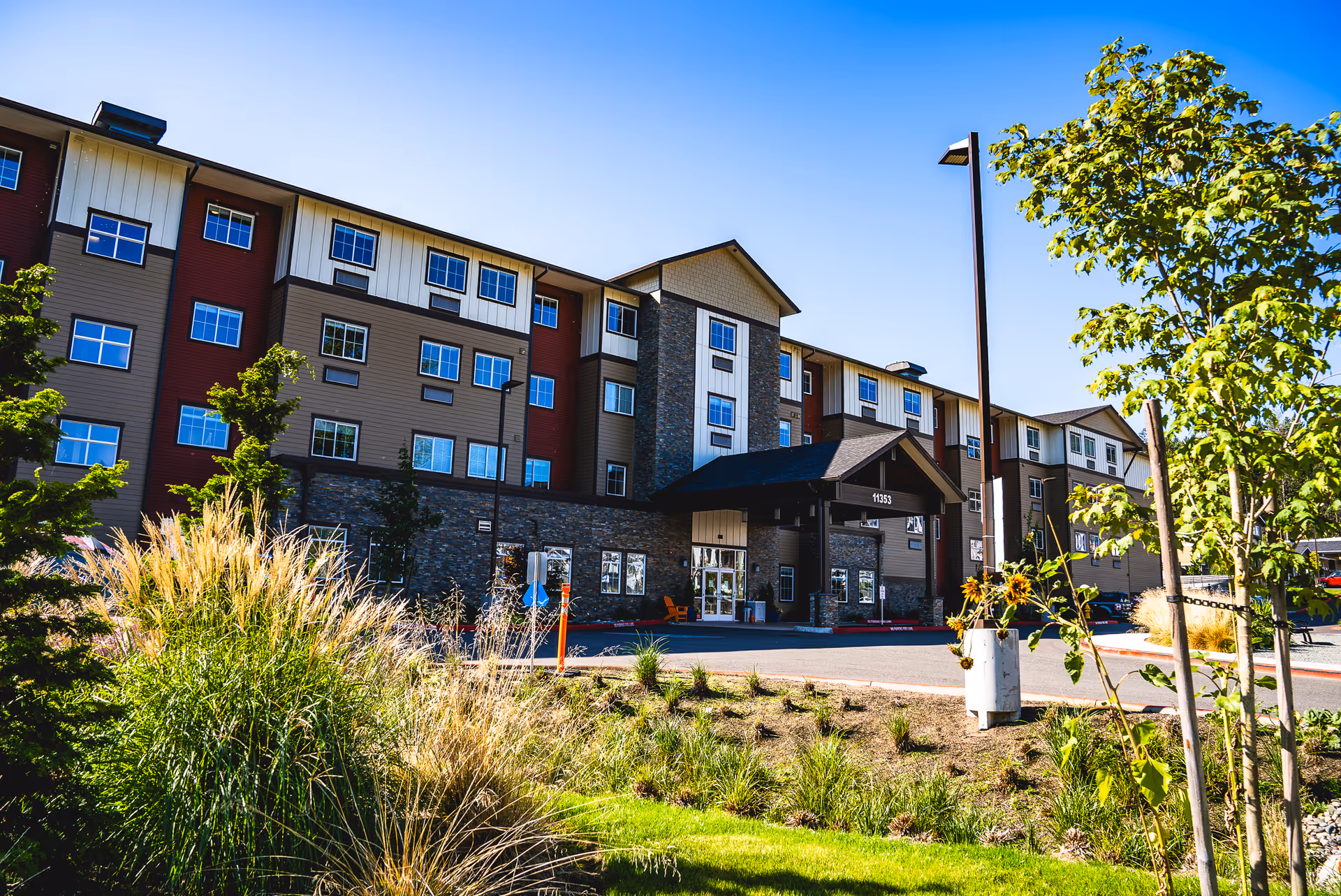 Four-story senior living building with a covered entrance, landscaped grounds, and a clear blue sky.