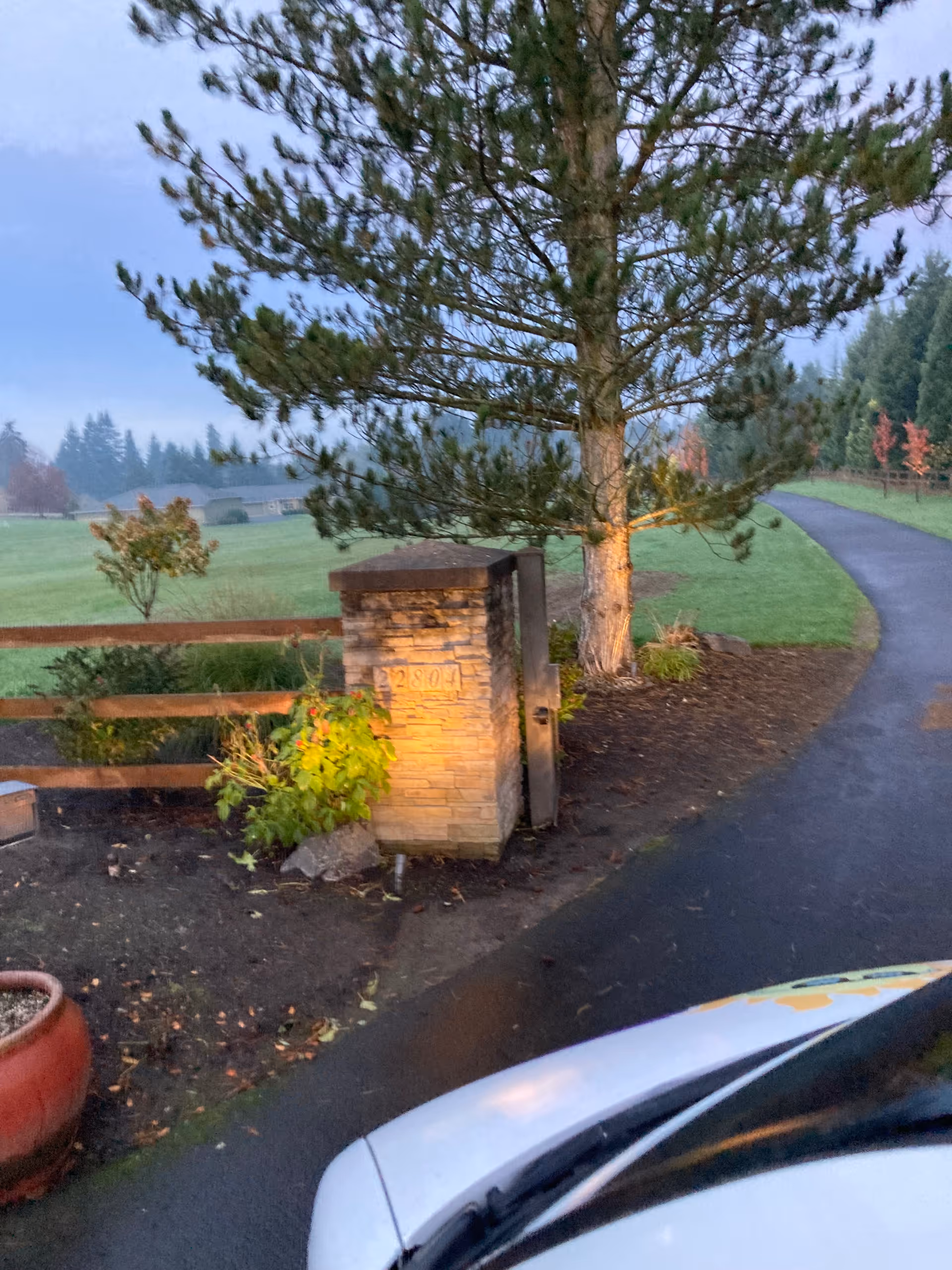 Paved driveway curves past a stone pillar, large tree, and landscaped lawn at the entrance.