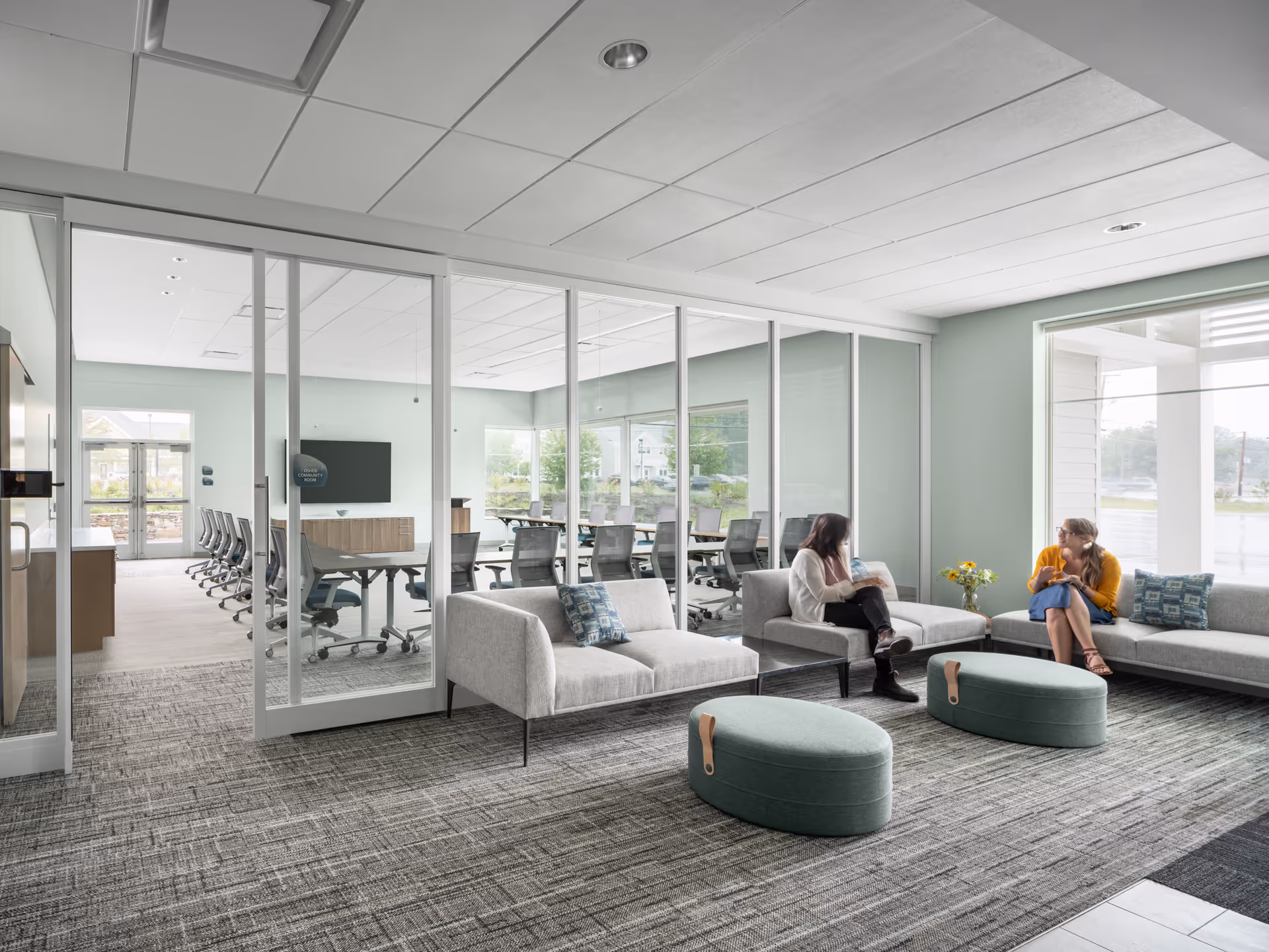 Modern lounge area with glass-walled conference room, couches, ottomans, and two people conversing.