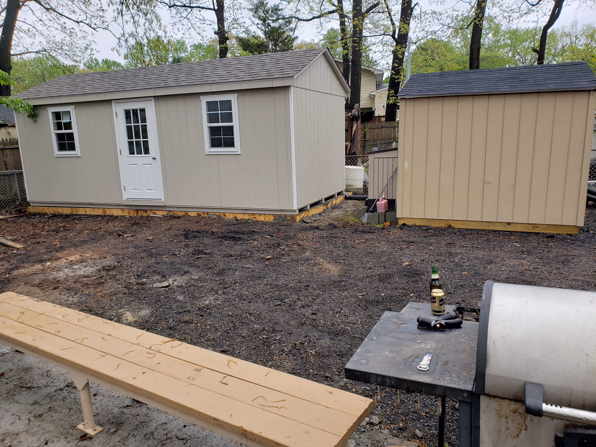 Two beige sheds in a muddy backyard with a grill table.