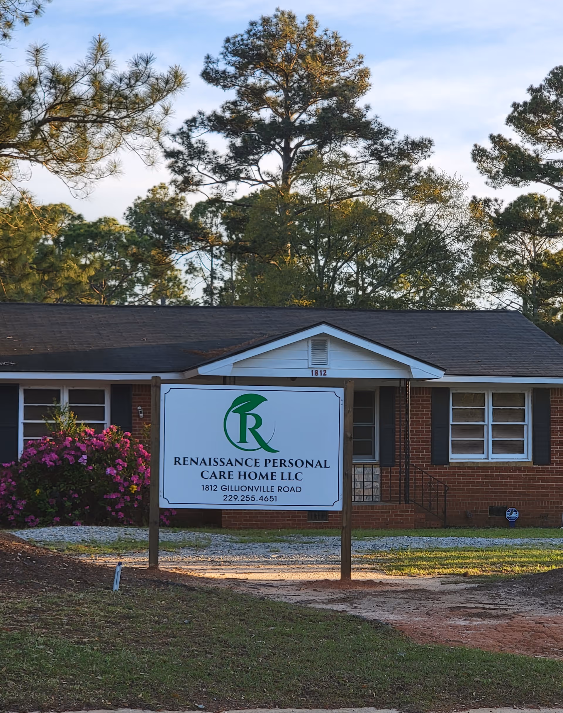 Exterior view of a single-story brick building with a dark roof, surrounded by trees and bushes with pink flowers. In front of the building is a large sign that reads 'Renaissance Personal Care Home LLC, 1812 Gillionville Road, 229.255.4651'.