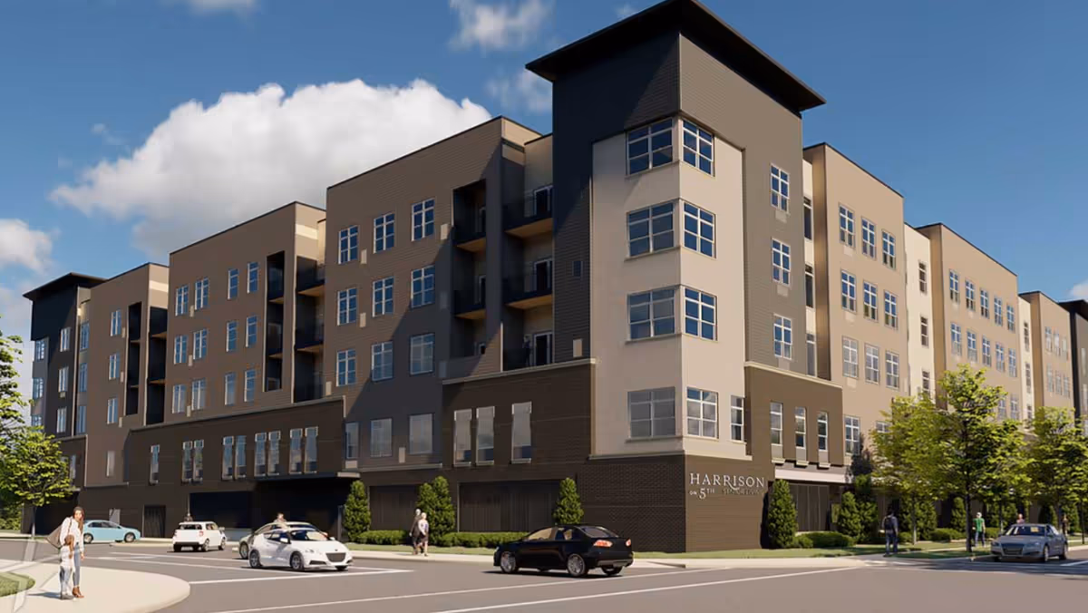 Exterior view of a modern multi-story senior living facility named Harrison on 5th by Senior Star, with several cars on the street and people walking on the sidewalks under a blue sky with some clouds.