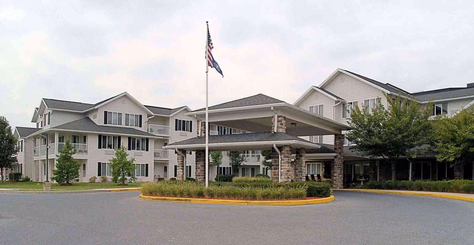Front entrance of a multi-story senior living facility with a covered porte-cochère, flagpole, and landscaped circular drive.