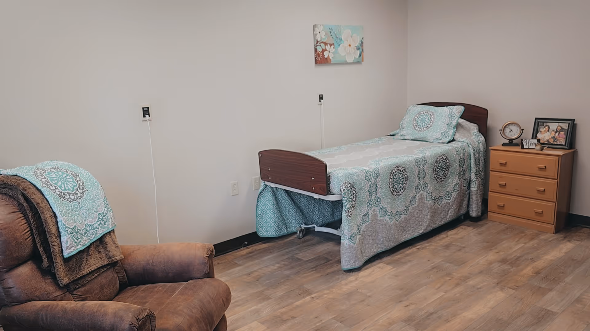 A furnished senior living bedroom with an adjustable hospital bed, a recliner, and a wooden dresser.