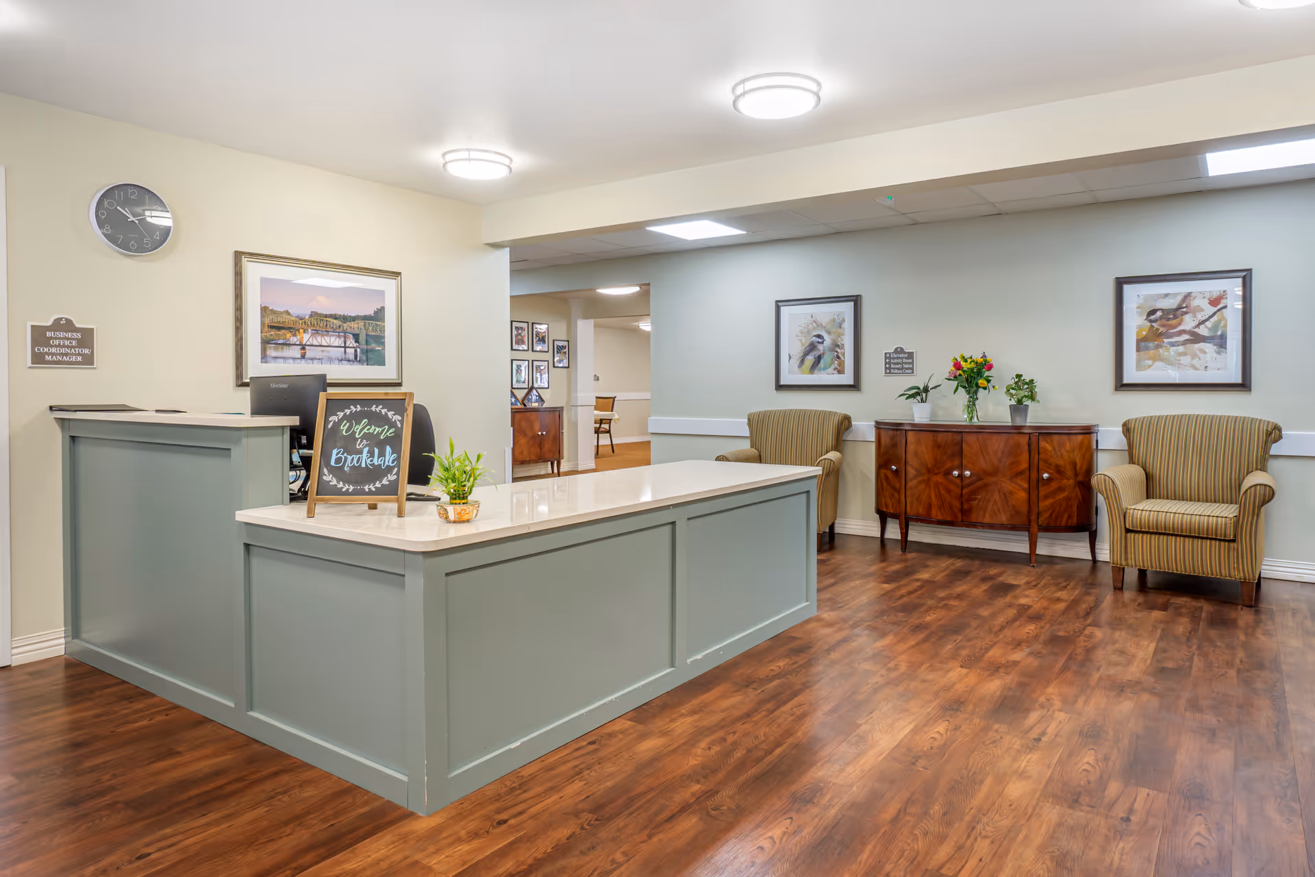 Reception area with a light green front desk featuring a small chalkboard sign that says 'Welcome to Brookdale.' Behind the desk is a computer and a framed landscape picture on the wall. To the right, there are two striped armchairs with a wooden cabinet between them, decorated with potted plants and a vase of flowers. The floor is wood, and the walls are painted light colors with framed bird artwork. Ceiling lights illuminate the space.