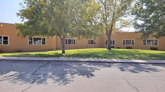 Single-story tan building with multiple windows, trees and a grassy lawn facing a street.