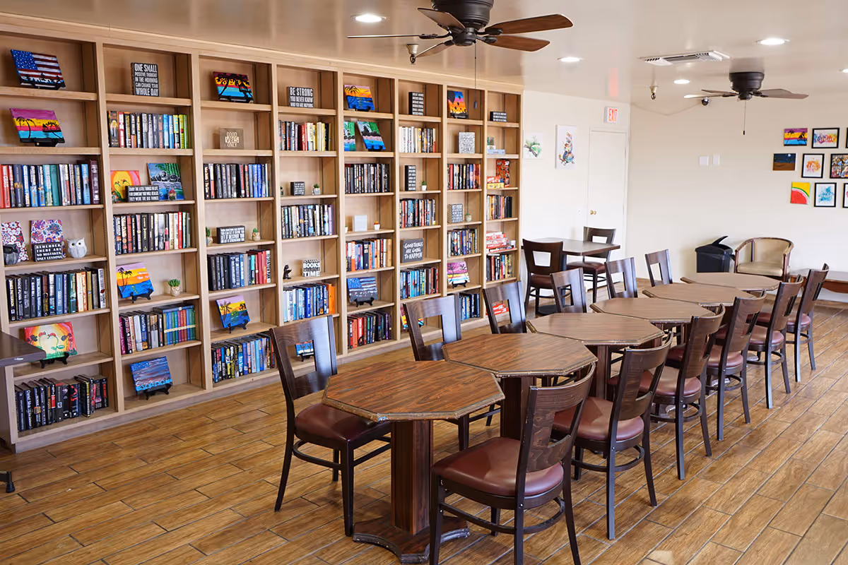 Communal activity room with wooden bookshelves along the wall and rows of wooden tables and chairs.