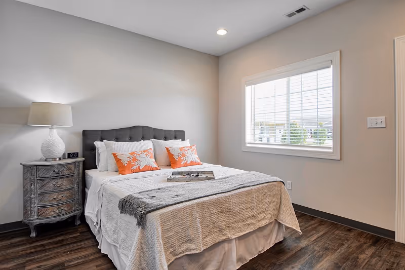 A neatly made bedroom featuring a bed, nightstand, lamp, and window.