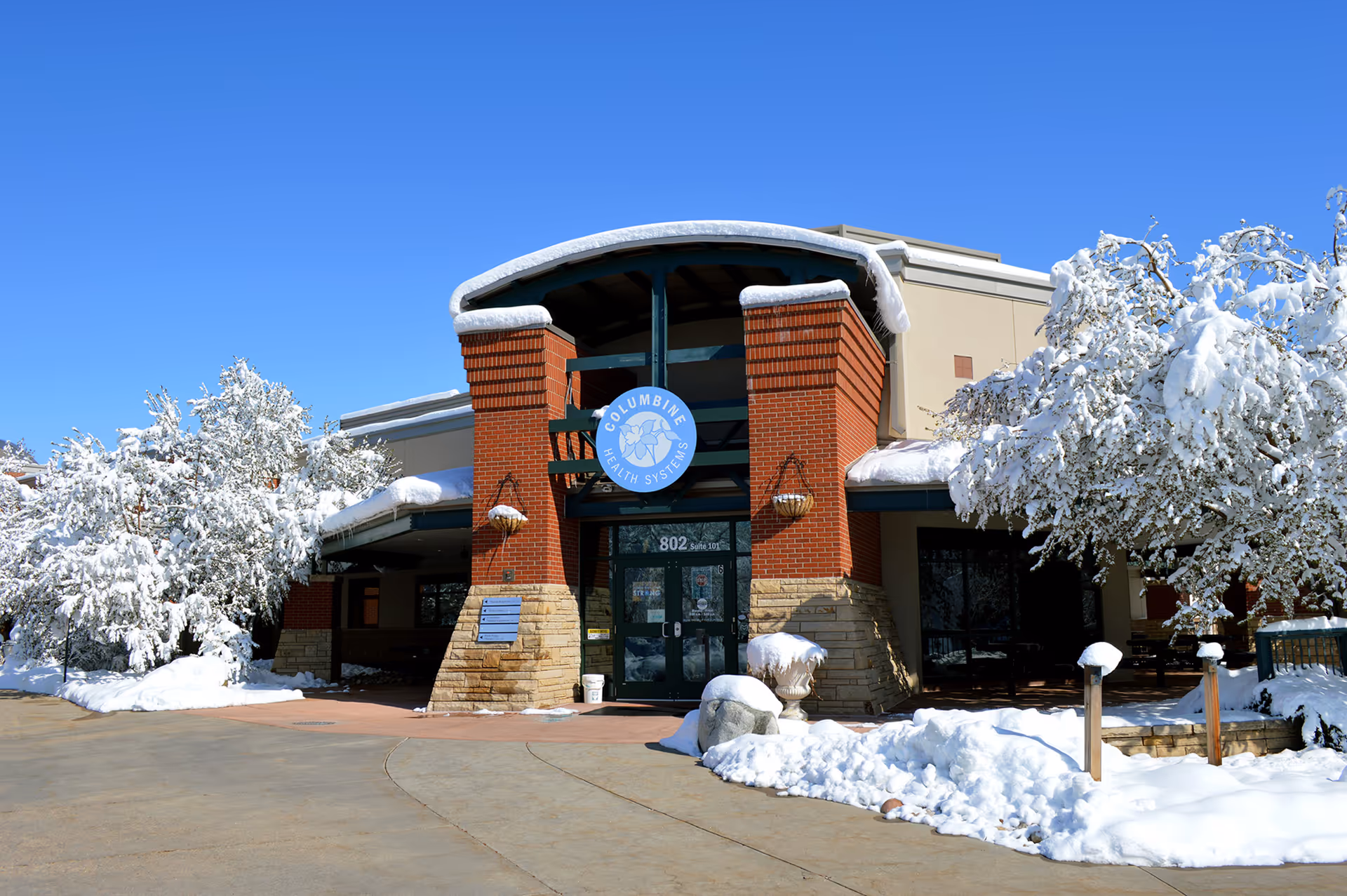 Snow-covered building entrance of Columbine Health Systems featuring brick columns and circular logo.