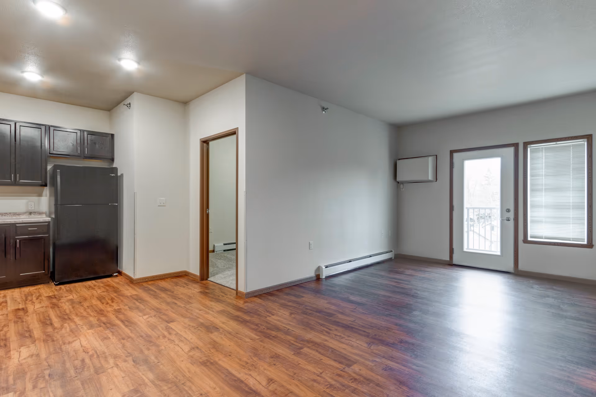 Empty apartment interior with wood flooring, a small kitchen area with dark cabinets and a black refrigerator on the left, a doorway leading to another room in the center, and a glass door with a window on the right letting in natural light.