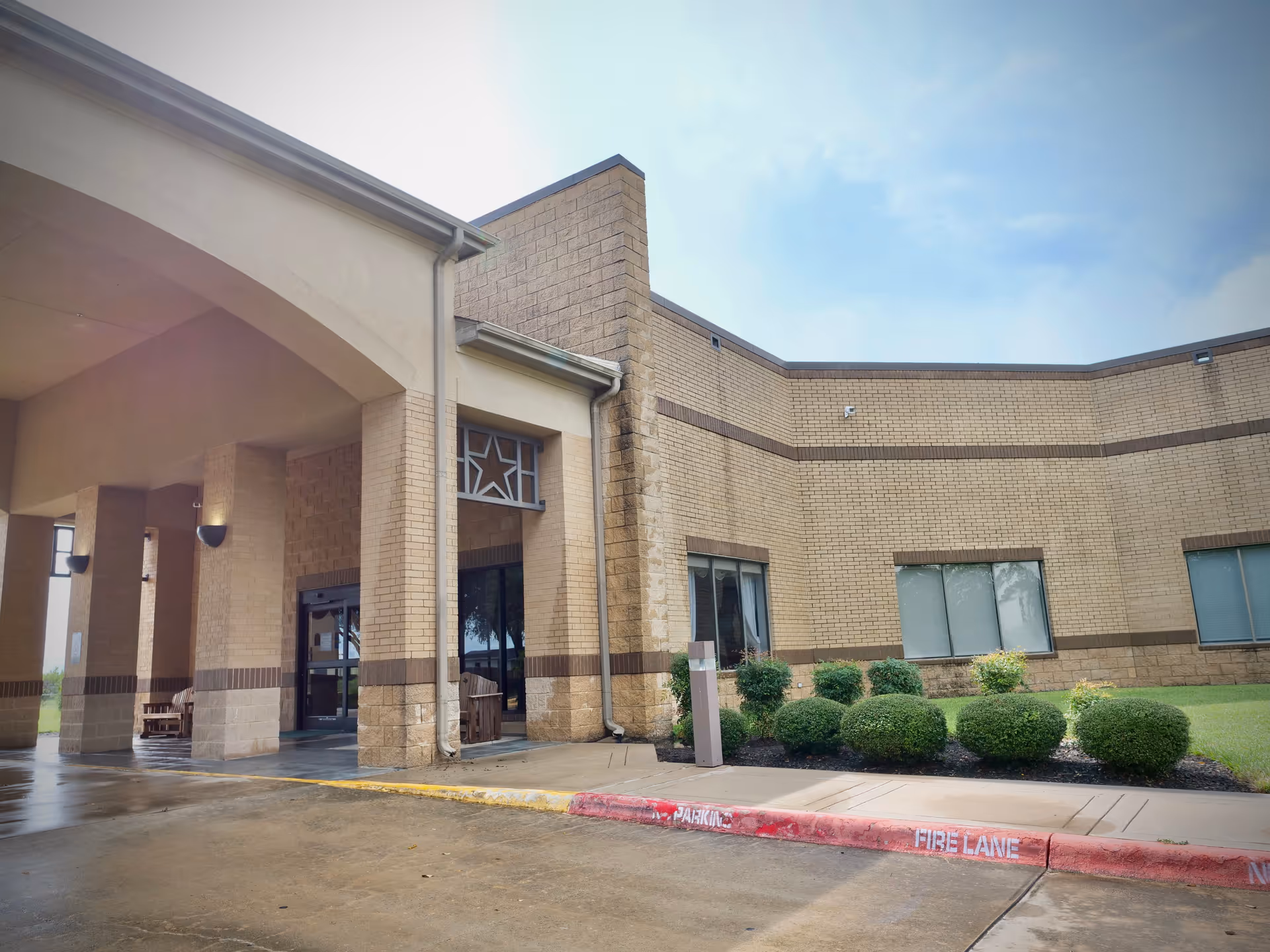 Exterior view of Twin Pines Nursing & Rehab Center showing the entrance with a covered drop-off area, beige brick walls, windows, and neatly trimmed bushes along the sidewalk. The sky is partly cloudy.
