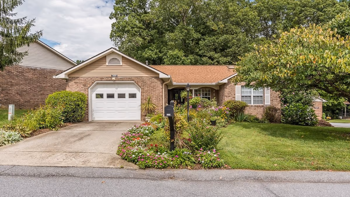 Single story brick senior living facility with a manicured lawn, colorful flower beds, and driveway.