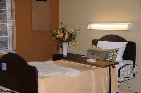 A hospital-style bedroom in a senior facility with a bed, overbed table, and flowers.
