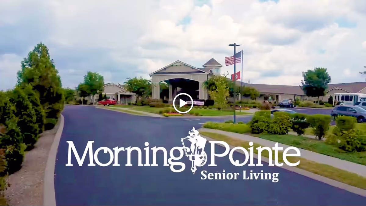 Front entrance of the Morning Pointe senior living facility with a driveway, landscaped grounds, and flagpoles.