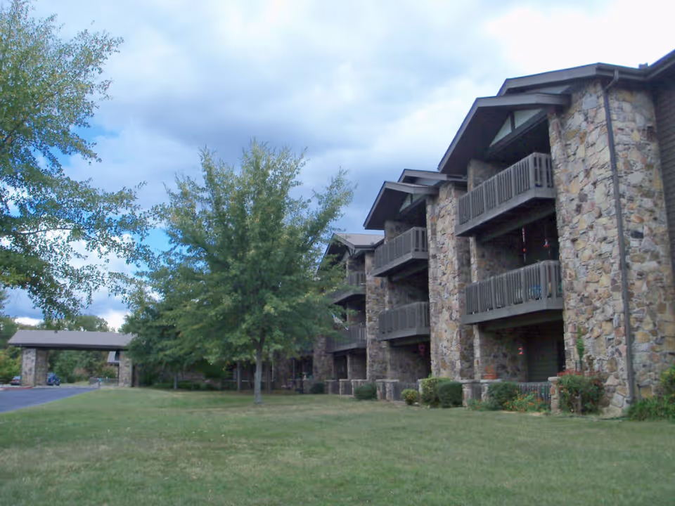 Stone-faced multi-story senior living building with balconies, lawn and trees under a cloudy sky.