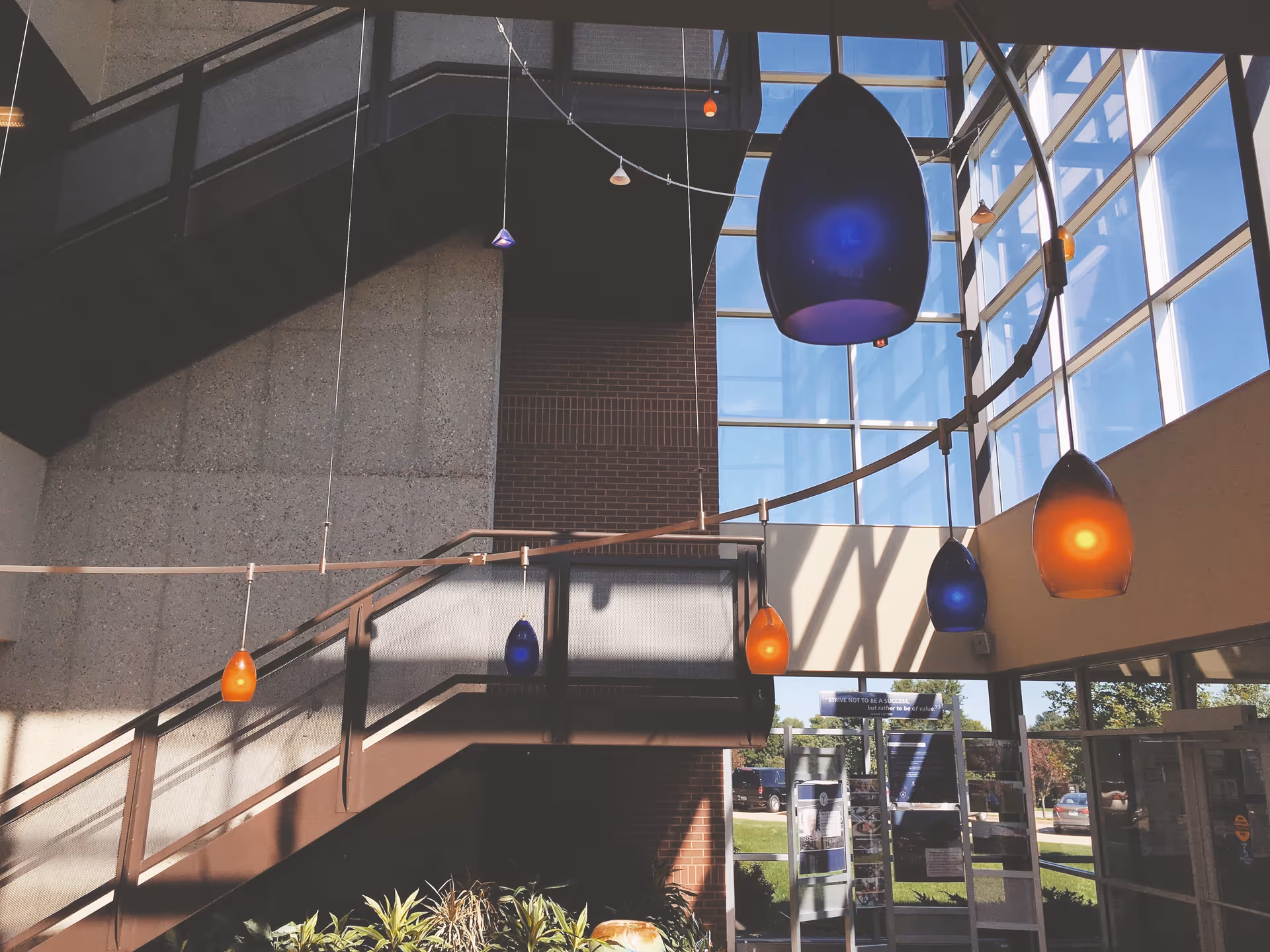 Sunlit interior atrium with a staircase, brick wall, and colorful hanging pendant lights.