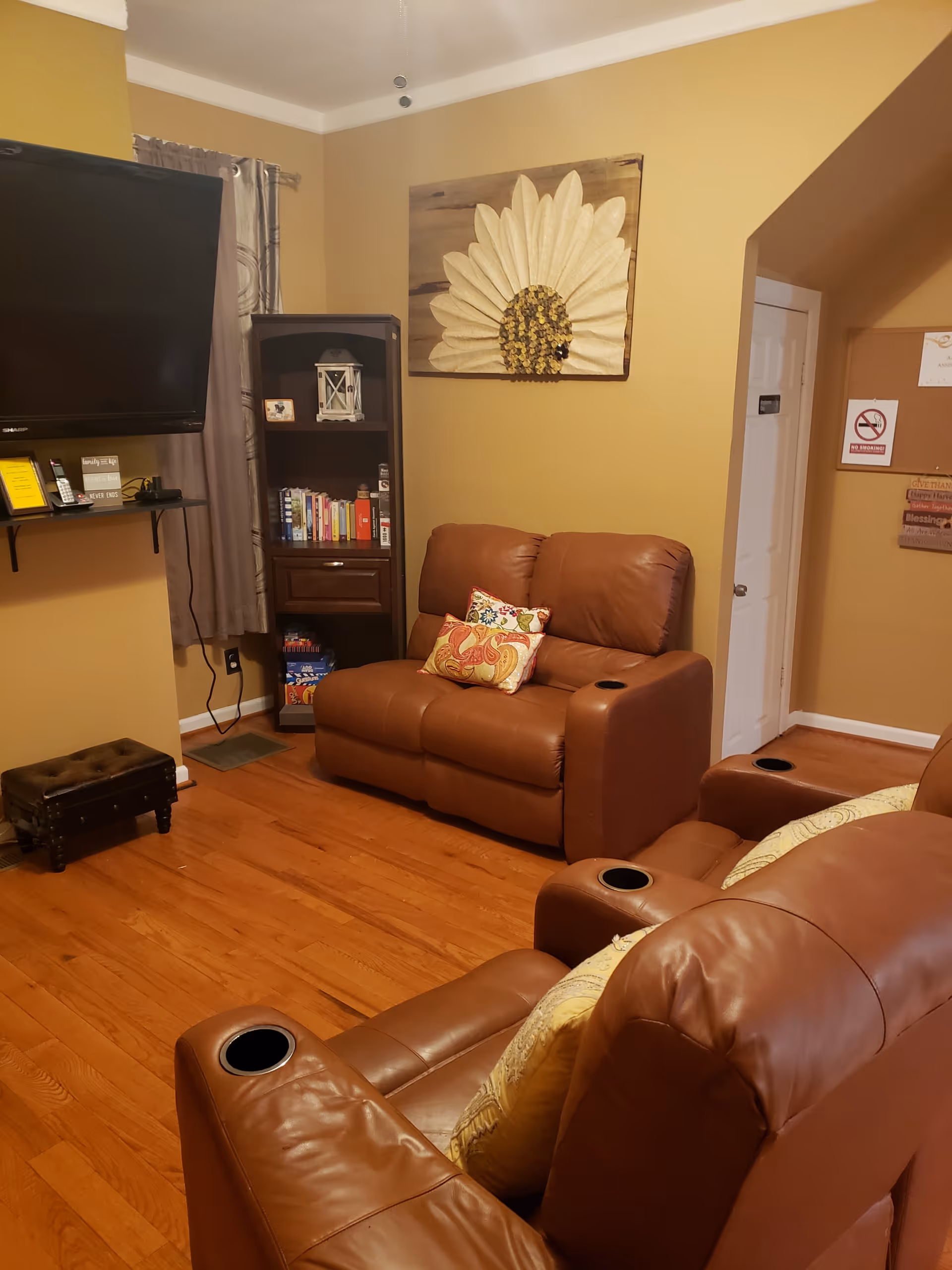 A cozy living room with two brown leather recliners and a matching loveseat, each with decorative pillows. A large flat-screen TV is mounted on the wall above a small shelf holding a phone and framed items. A dark wooden bookshelf filled with books and decorative items stands in the corner. The walls are painted a warm beige color and adorned with a large flower artwork. Hardwood floors complete the room.