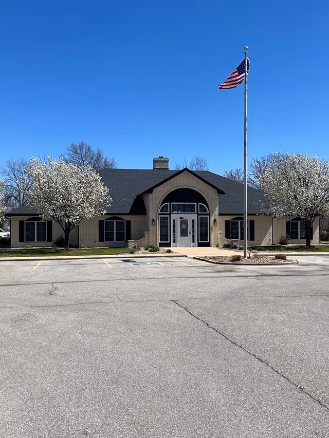 Exterior view of Oswego Home Place Senior Living building with flagpole and blossoming trees