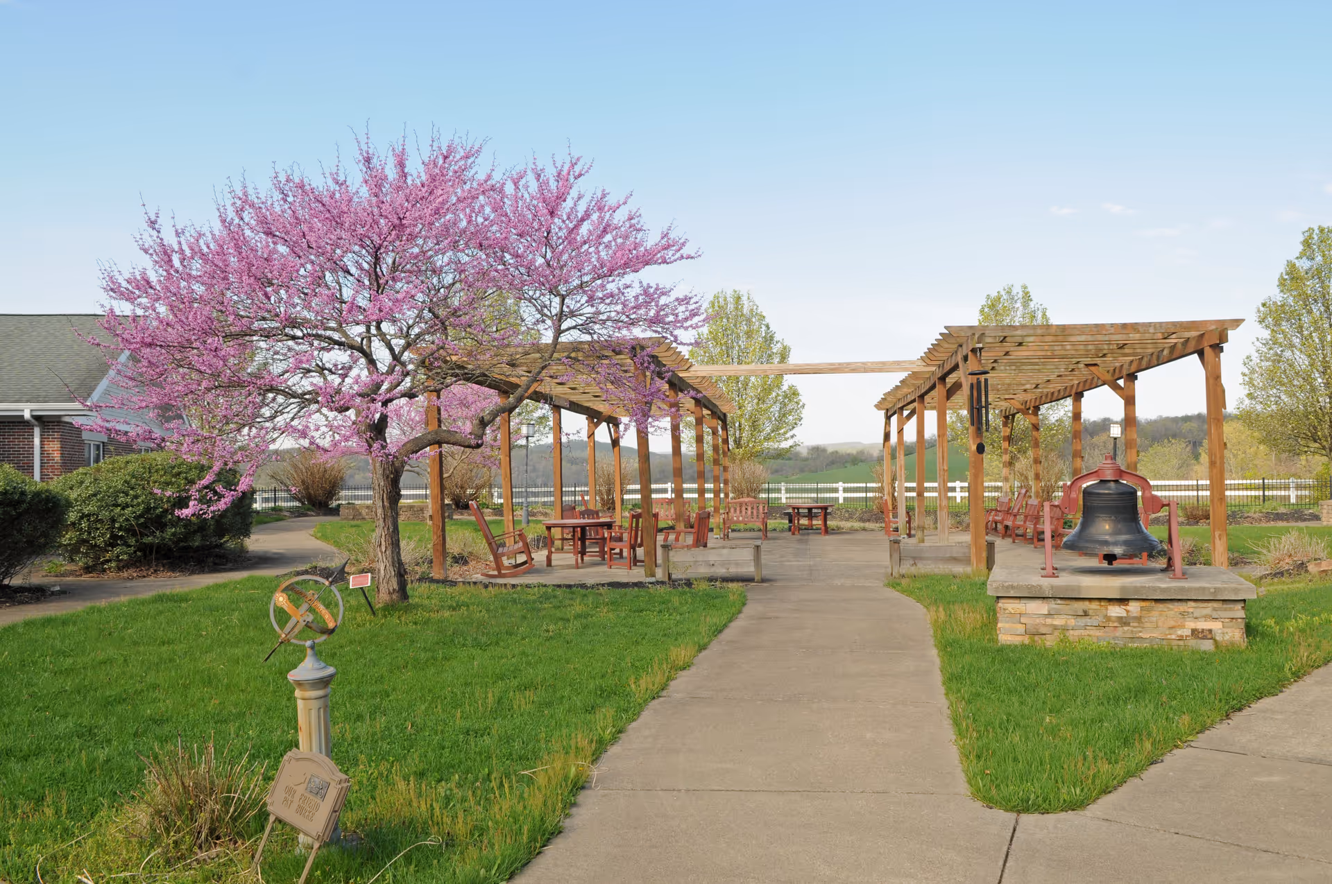 Outdoor garden area with a concrete pathway leading to wooden pergolas with benches and tables underneath. A tree with pink blossoms is on the left side, and a large bell mounted on a stone platform is on the right. Green grass and shrubs surround the area, with a clear blue sky in the background.