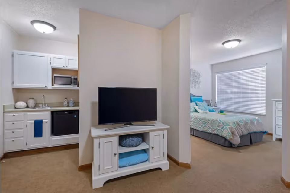 A cozy bedroom with a kitchenette featuring a small refrigerator, microwave, and a television on a white cabinet. The bedroom has a bed with decorative pillows and a window with blinds.