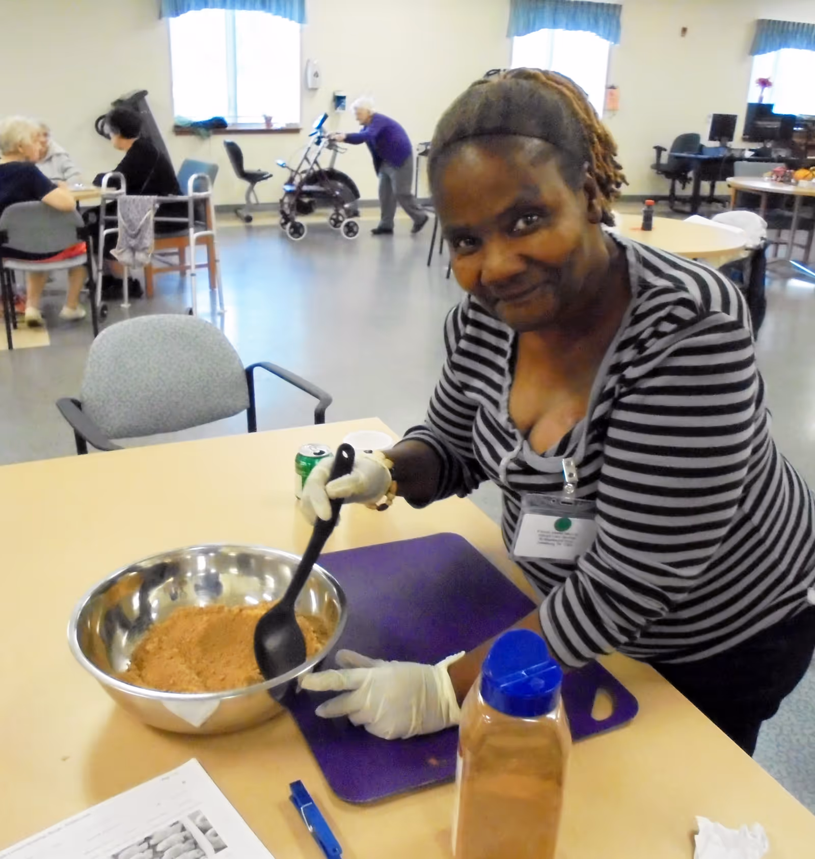 Caregiver wearing gloves prepares ingredients at a table in a senior living facility kitchen.