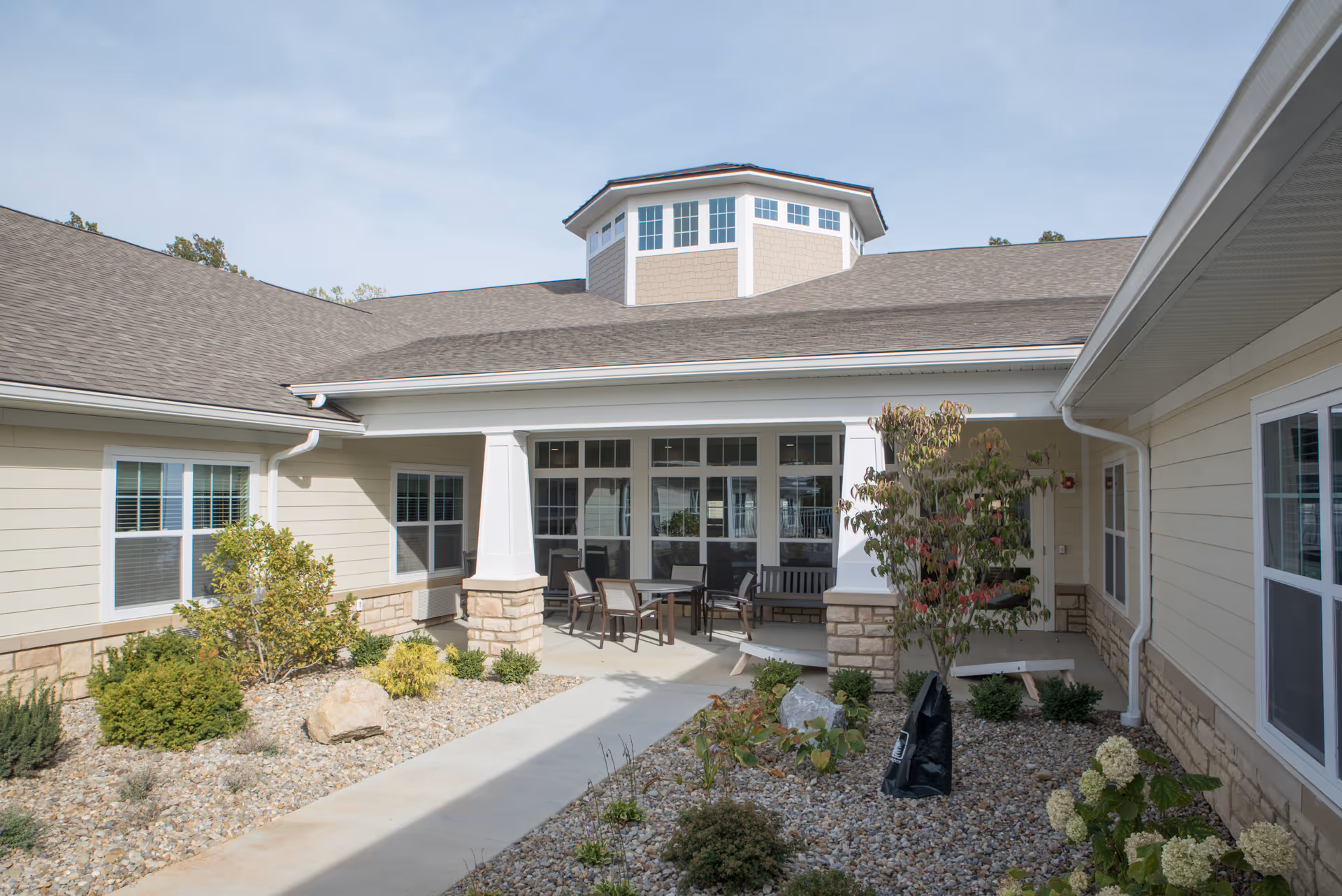 Sunny courtyard with a paved walkway, outdoor seating, windows and landscaped rock beds between single-story beige buildings.