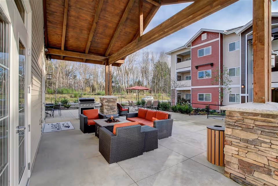 Outdoor covered patio area with a wooden roof structure, featuring a seating arrangement with black wicker furniture and red cushions around a fire pit. In the background, there is a barbecue grill, tables with chairs, and a red umbrella. A multi-story residential building with red and beige siding is visible, along with trees and a clear sky.