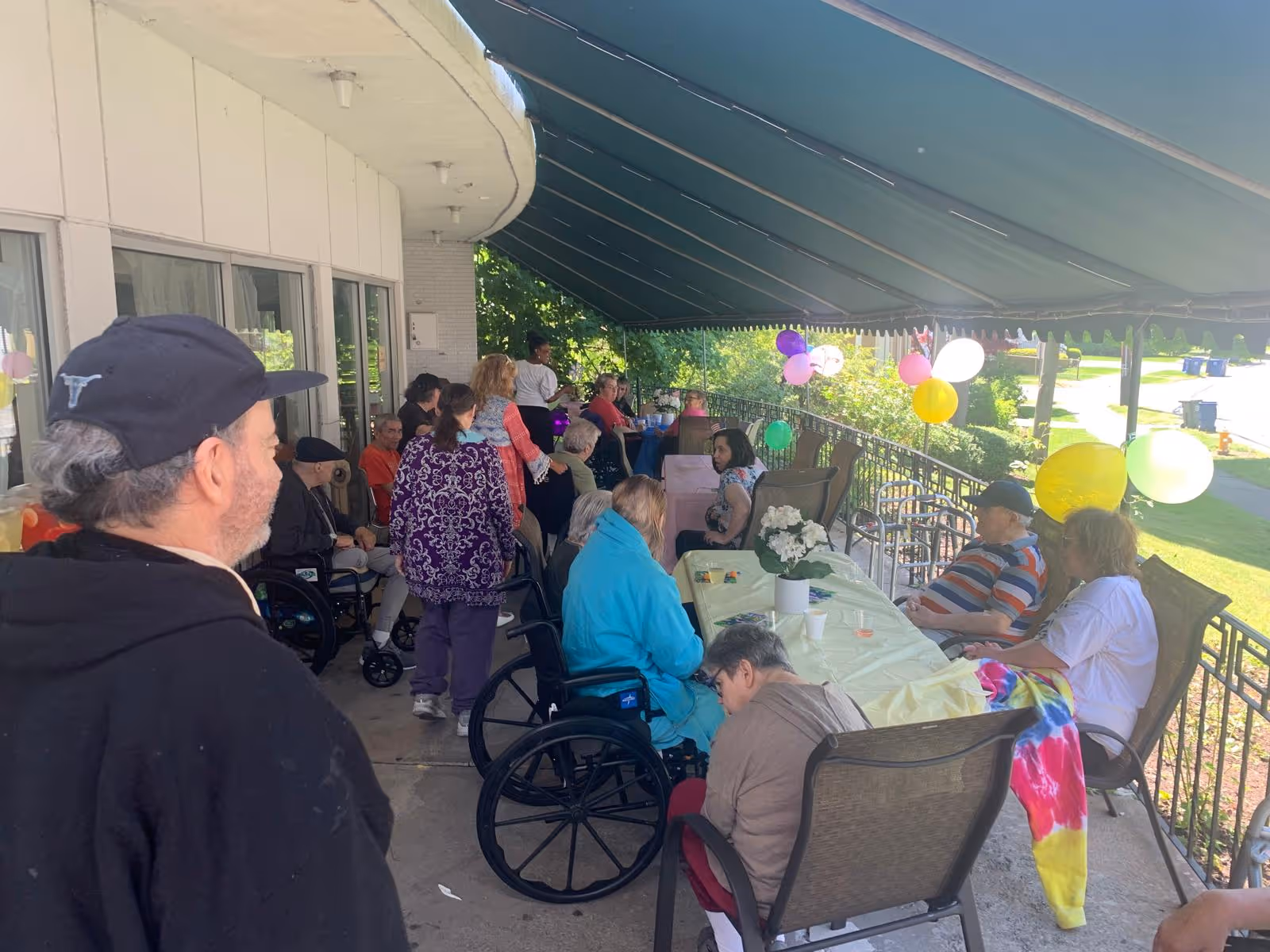 Elderly residents and caregivers gather on a shaded outdoor patio with balloons.
