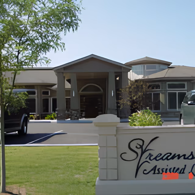 Front exterior view of Streamside Assisted Living facility showing the entrance with a covered porch, surrounded by windows and landscaping, with a sign in the foreground displaying the facility's name.