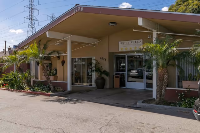 Front entrance of Rose Villa Healthcare Center with a sloped canopy, potted palms, glass doors, and the address number 9028.