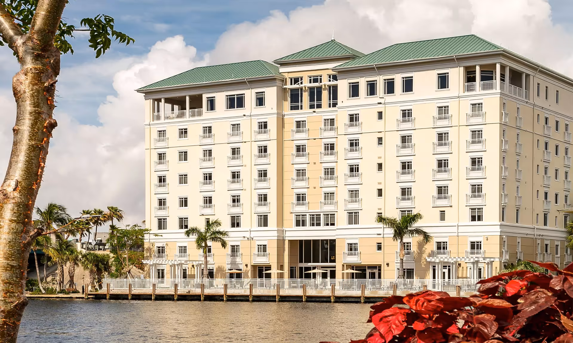 Seven-story beige senior living building with a green roof overlooking a waterfront with palm trees and a dock.