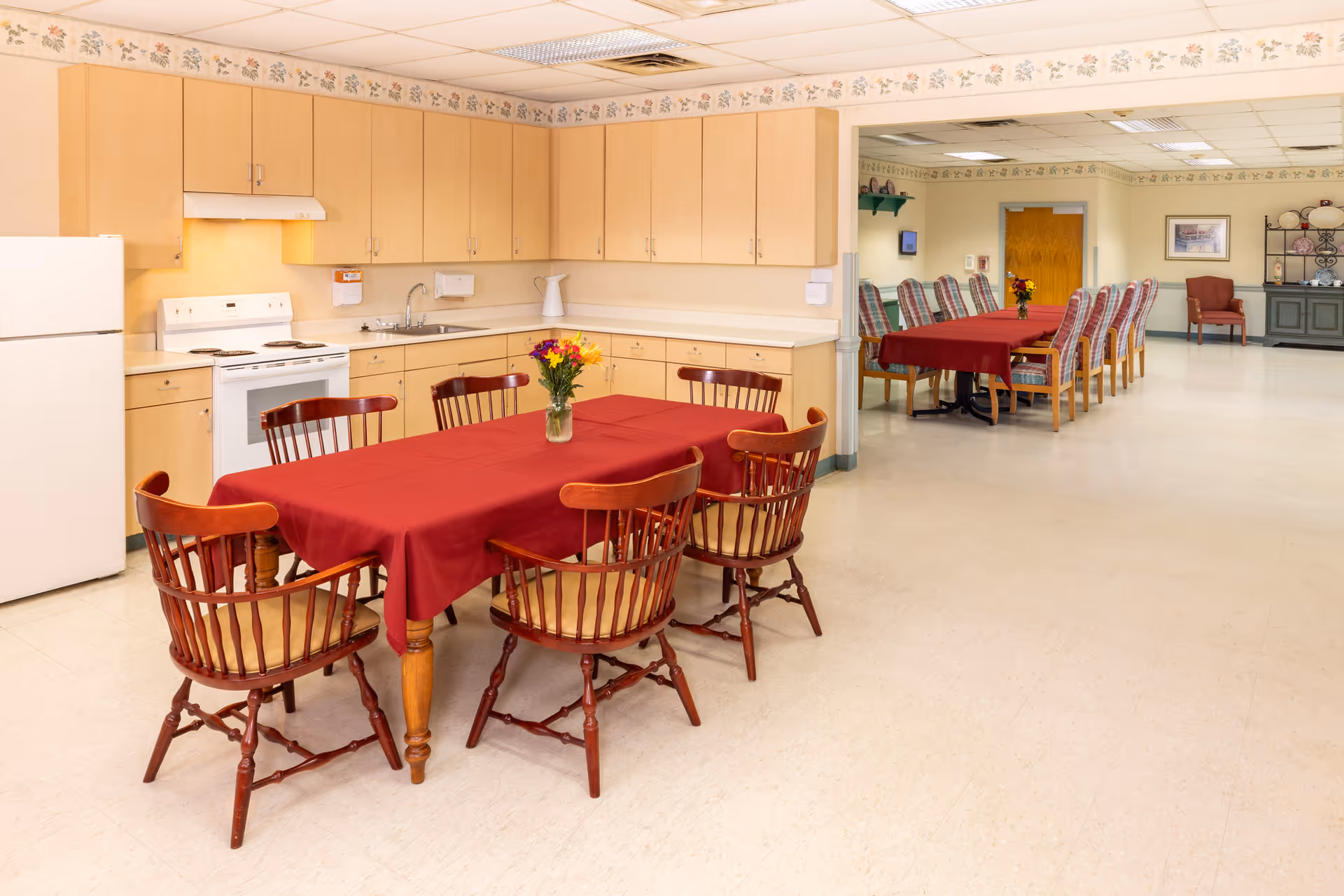 A communal dining area with red tablecloths, wooden chairs, and a kitchen along the left wall.