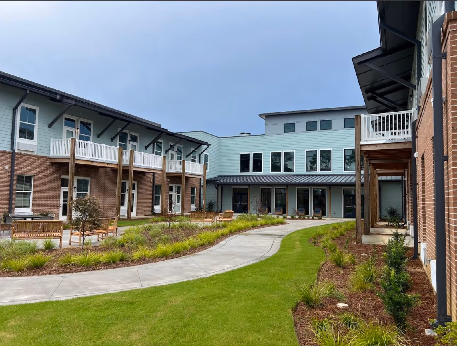 Outdoor courtyard area at The Marsh at Carolina Park featuring a curved concrete walkway, green grass, landscaped plants, wooden benches, and a two-story building with brick and light blue siding, balconies, and multiple windows under a cloudy sky.