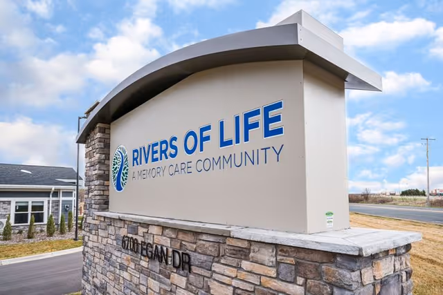 Outdoor stone and concrete sign for Rivers of Life, a memory care community, located at 6700 Egan Dr, with a partly cloudy sky in the background.