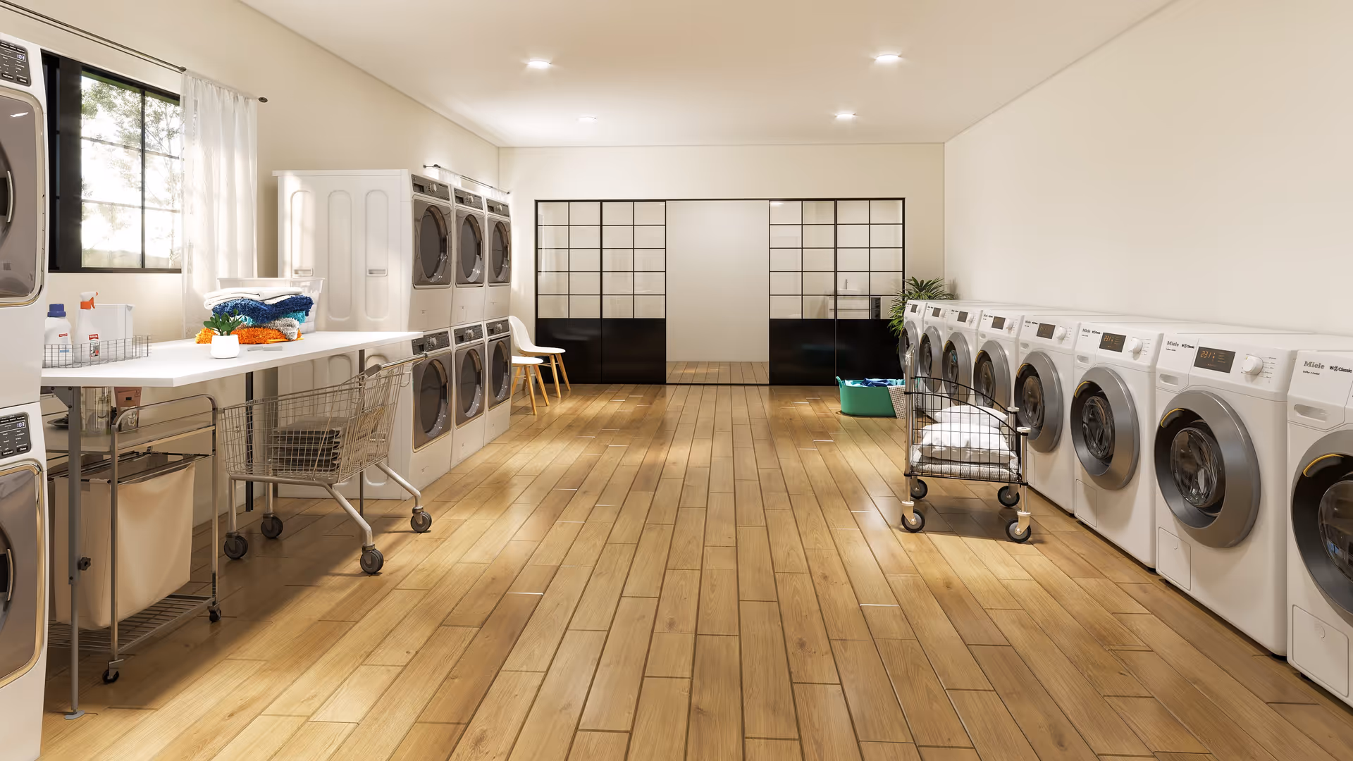 Spacious, well-lit communal laundry room with rows of front-loading washers and dryers, folding tables, carts, and wooden floors.