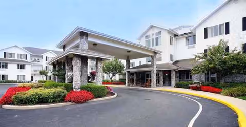 Senior living facility exterior with covered entrance, stone columns, curved driveway, and landscaped shrubs.