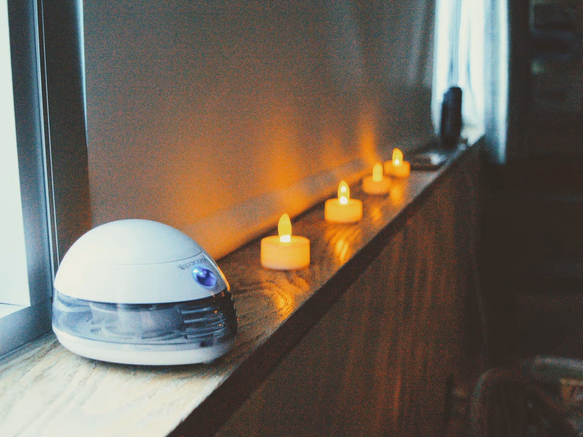 Row of lit candles on a wooden windowsill beside a white round device.