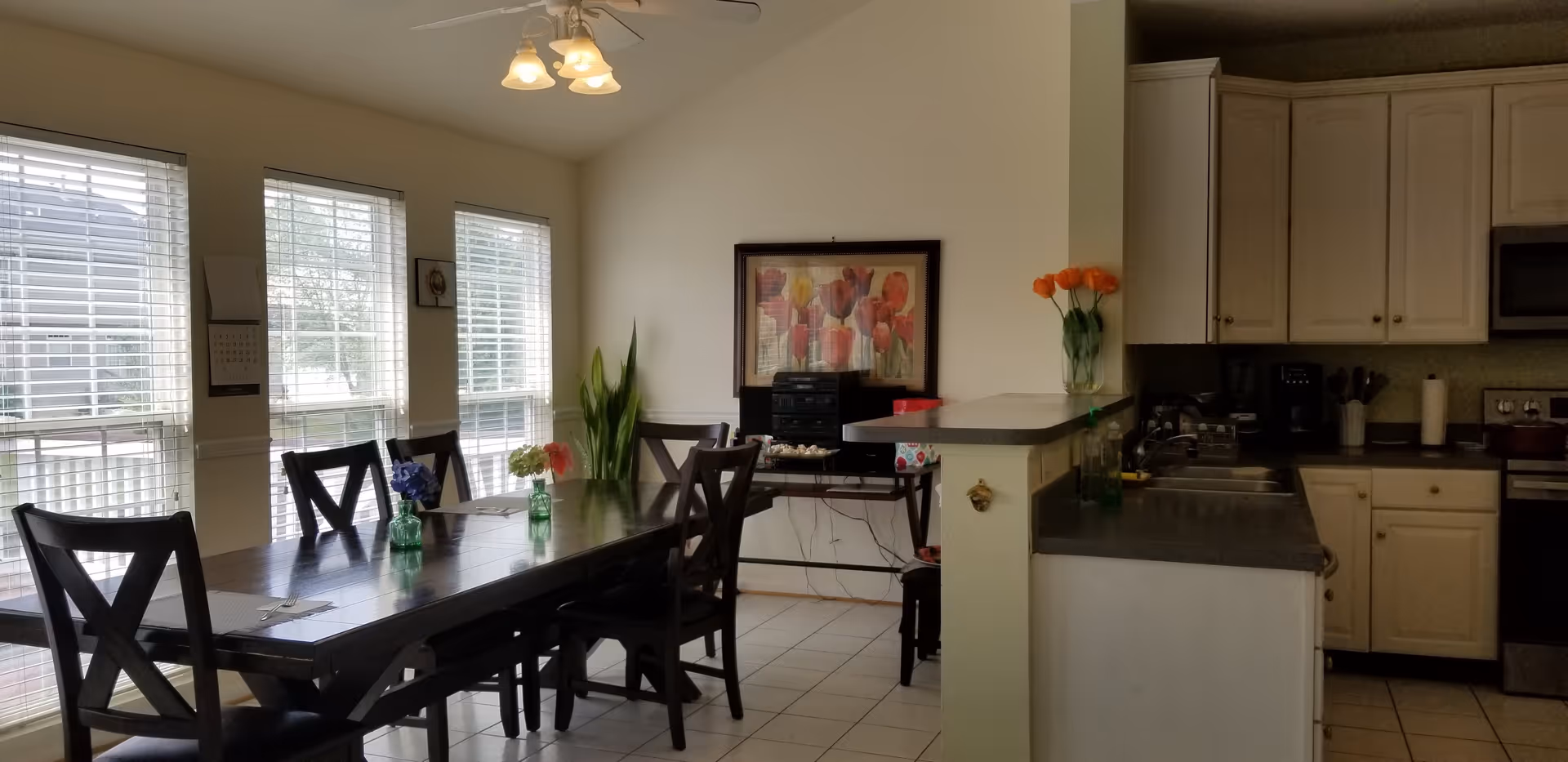 A bright dining area with a long dark wooden table surrounded by six matching chairs. The table has small vases with colorful flowers. Large windows with white blinds let in natural light. Adjacent to the dining area is a kitchen with white cabinets, a dark countertop, and stainless steel appliances. A framed floral painting and a tall green plant decorate the wall near the dining table.