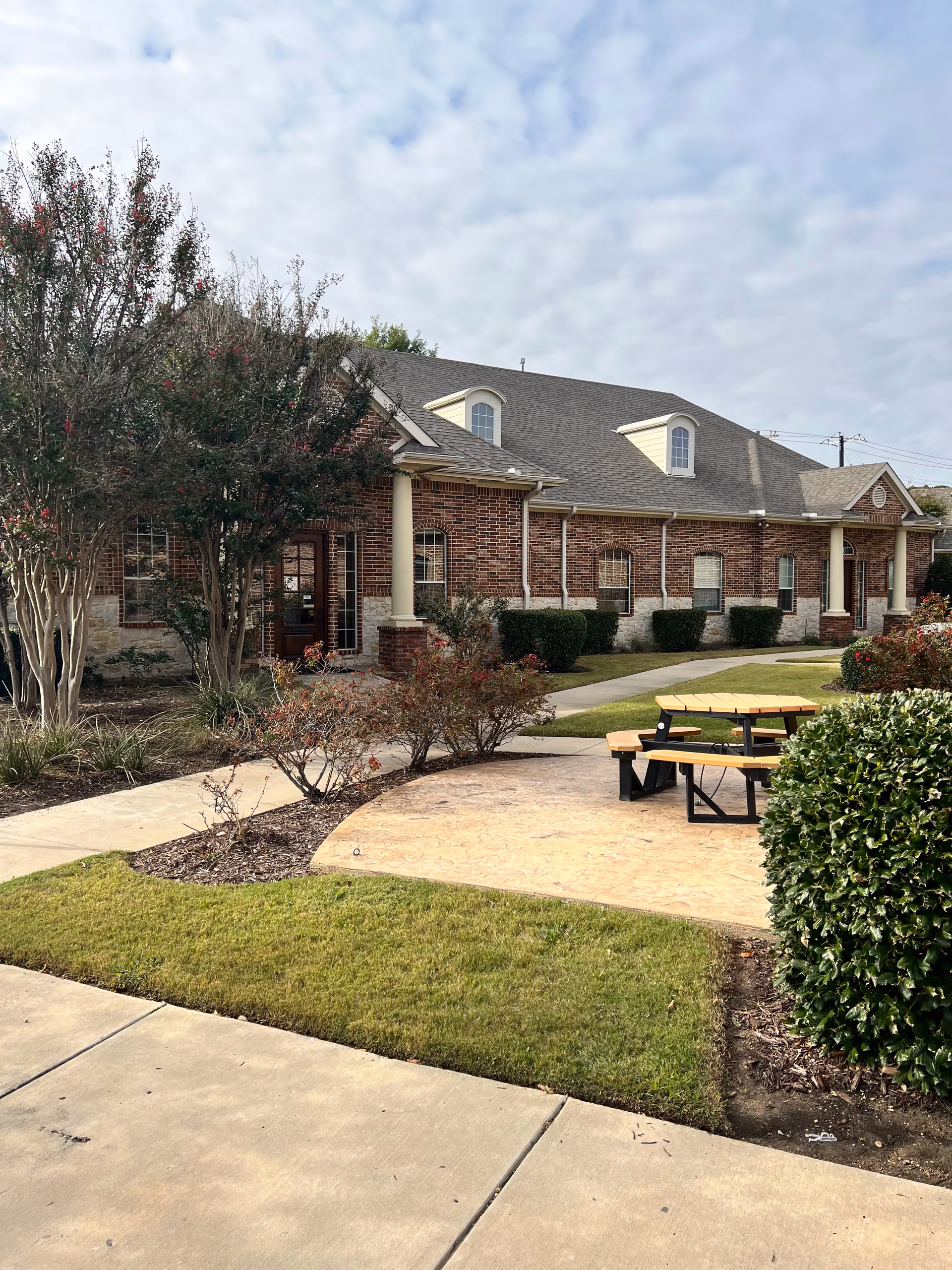 Brick senior living facility exterior with walkway, columns, and a picnic table.