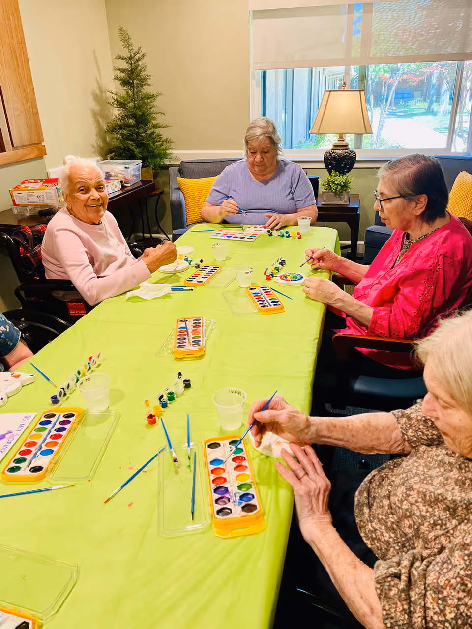 Four elderly women sitting around a table covered with a green tablecloth, engaging in a watercolor painting activity with paint palettes, brushes, and small cups of water. The room has a window with a view of greenery outside, a lamp on a side table, and a small potted plant.