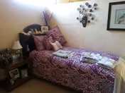 A cozy bedroom with a patterned purple bedspread, pillows, and framed wall decor.
