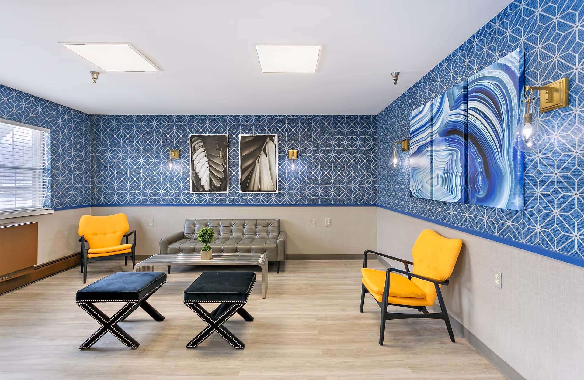 A modern sitting area with blue patterned wallpaper and light wood flooring. The room features a gray tufted sofa, two yellow armchairs with black frames, and two black cushioned stools with nailhead trim. A rectangular coffee table with a small green plant is placed in front of the sofa. The walls are decorated with black and white framed photos and a three-panel blue abstract artwork. The room is lit by ceiling lights and wall sconces with exposed bulbs.