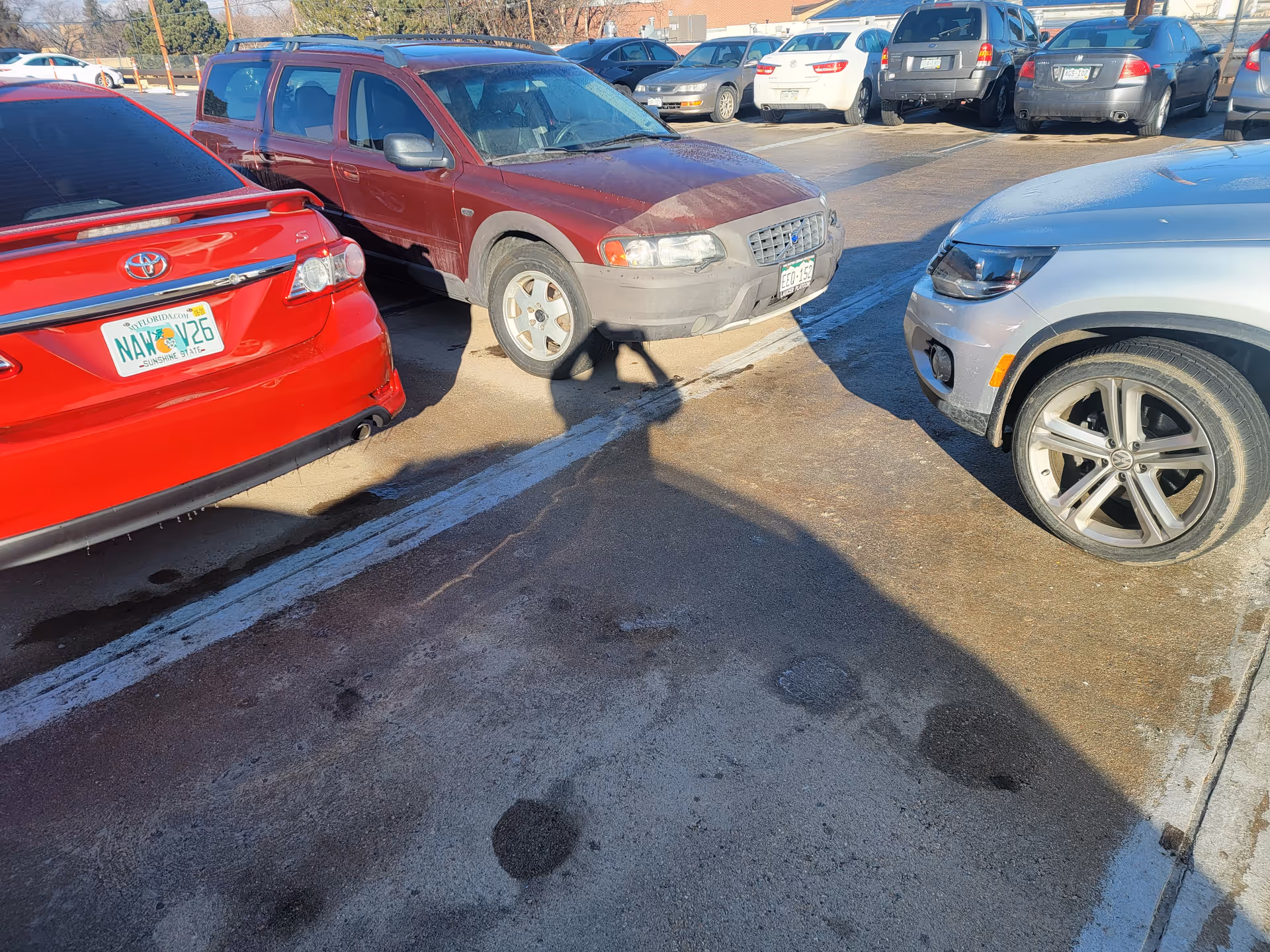 Sunlit parking lot with several cars and long shadows across the pavement.