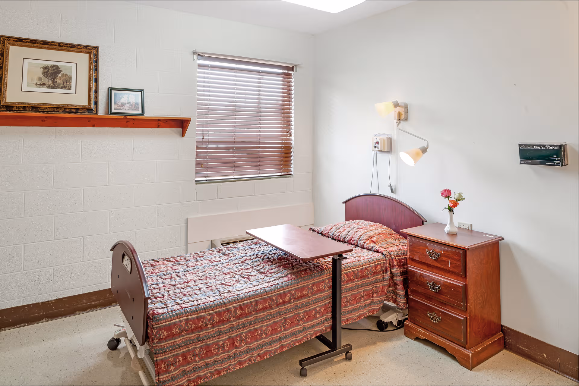 Bedroom in a senior living facility featuring a bed, dresser, nightstand, and window blinds.