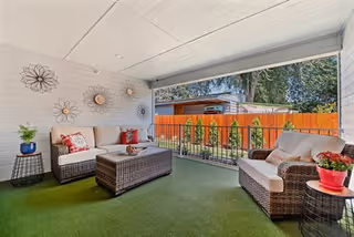 Covered outdoor patio area with artificial grass flooring, featuring two wicker sofas with cushions and pillows, a wicker coffee table, side tables with potted plants, decorative metal wall art, and a wooden fence with greenery in the background.