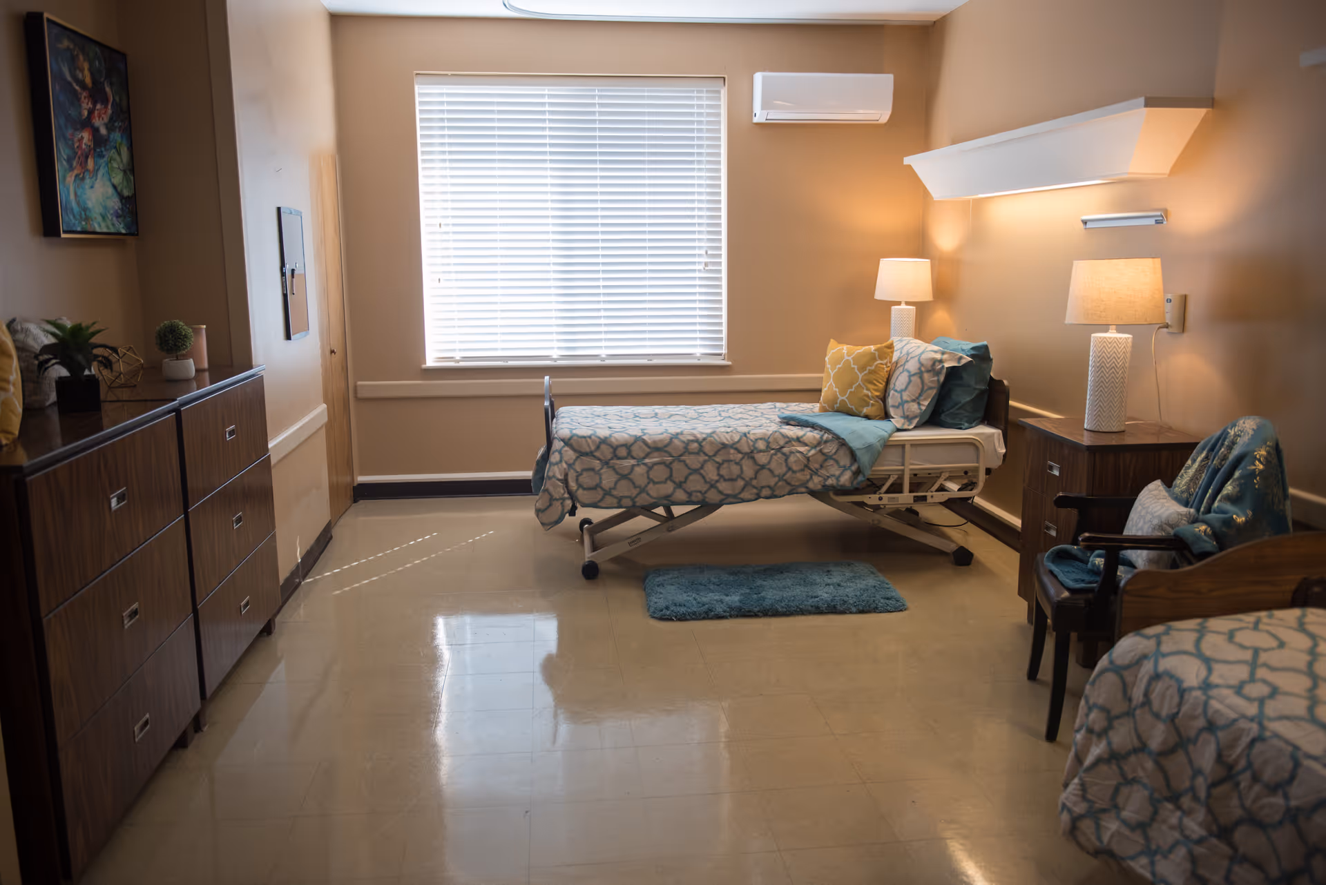 A clean beige patient room with two beds, nightstands, lamps, and a window.