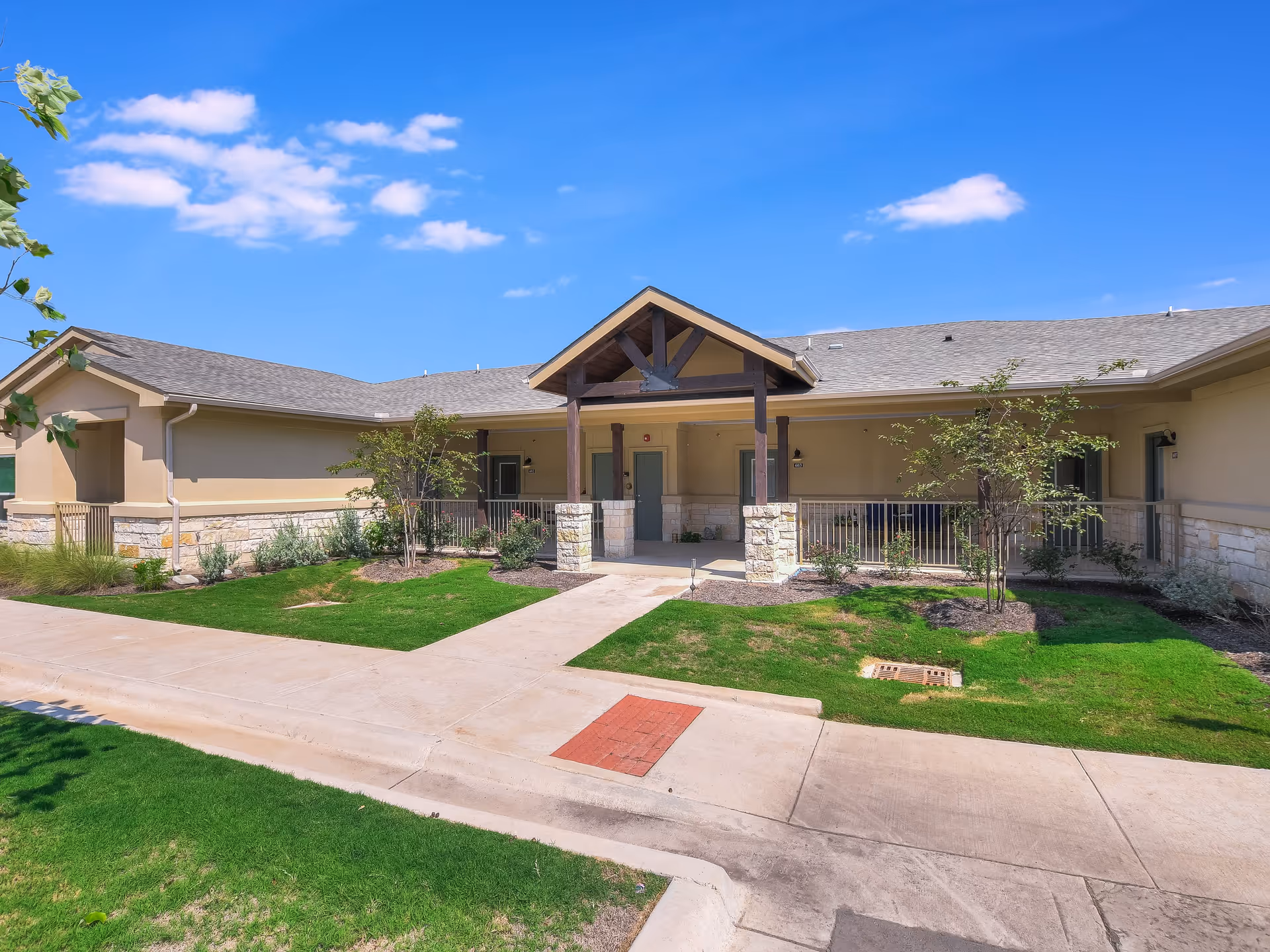 Front entrance of a single-story senior living building with a covered porch, walkway, and landscaped lawn under a blue sky.