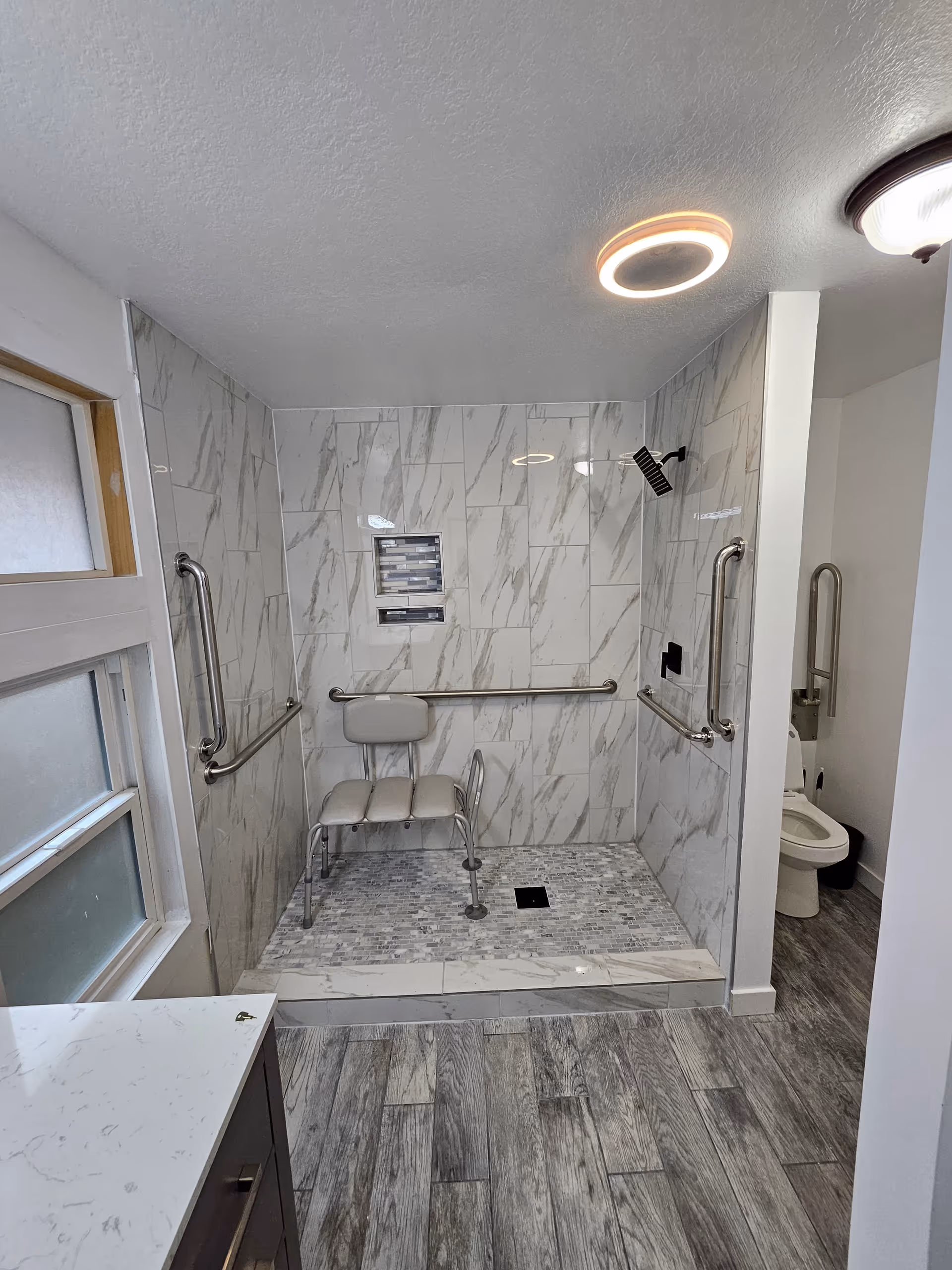 Accessible bathroom with marble tile shower, grab bars, built in seat, and visible toilet.