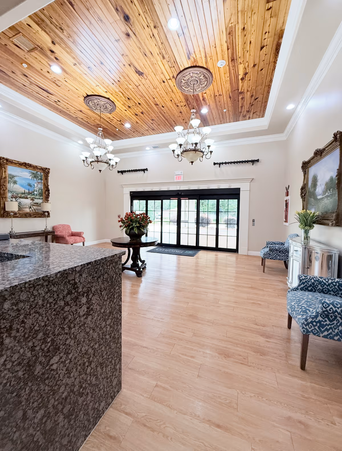 Spacious elegant lobby with a wooden coffered ceiling, chandeliers, seating, a central table with flowers, and glass entry doors.